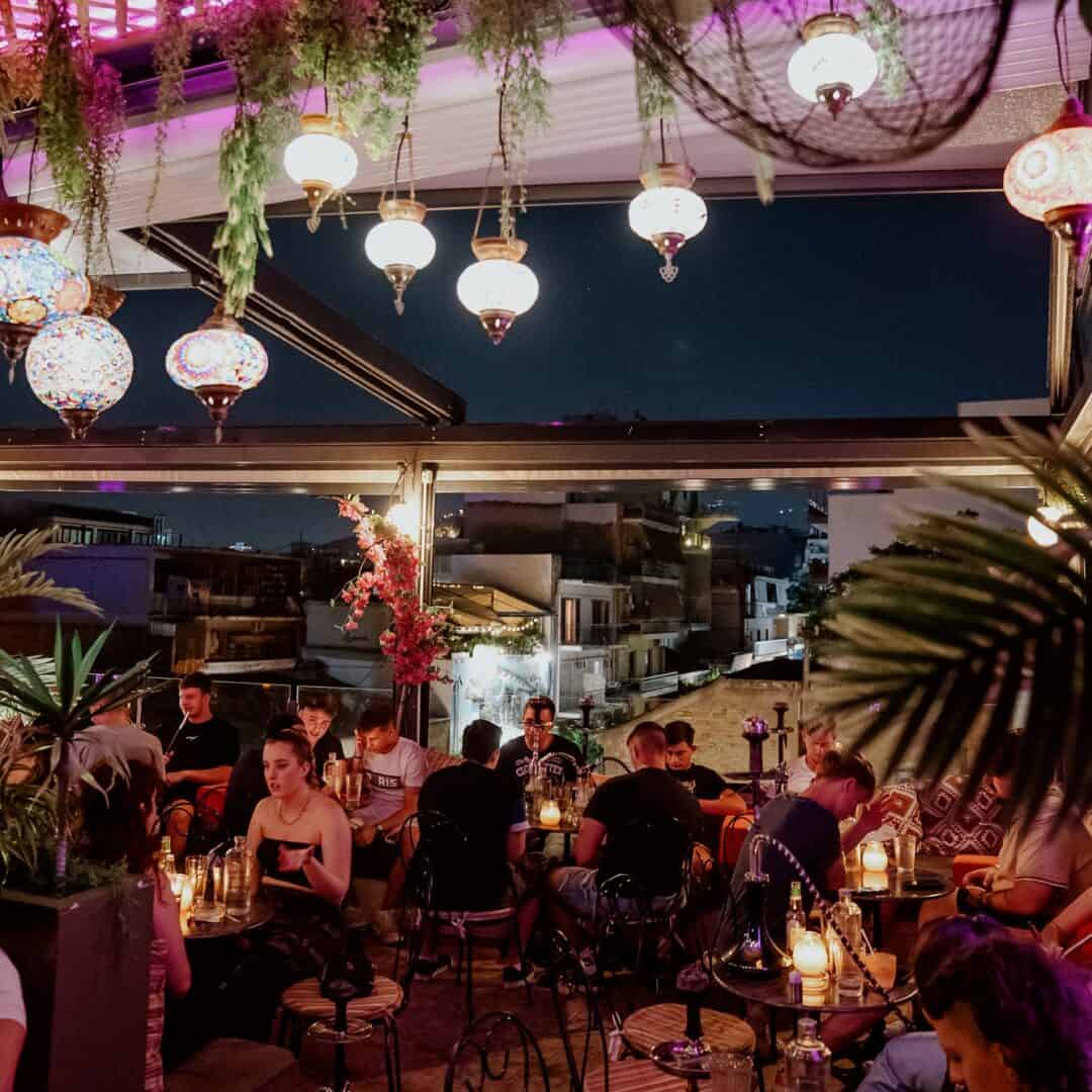 Vibrant rooftop lounge with hanging lanterns and guests enjoying shisha and drinks.